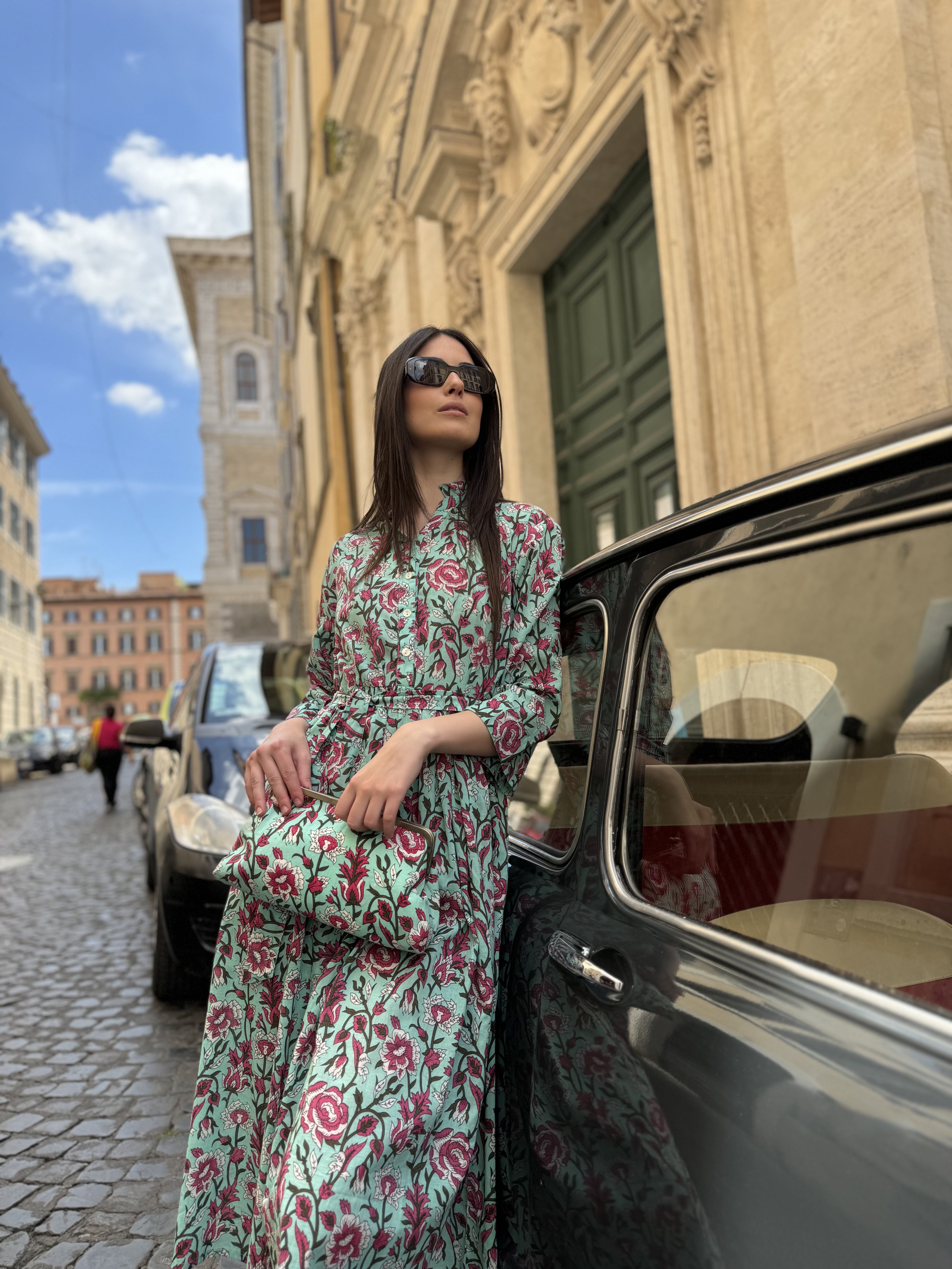 Donna elegante con abito floreale in mussola di cotone con borsetta abbinata appoggiata a un'auto d'epoca italiana in strada storica nuovissima collezione